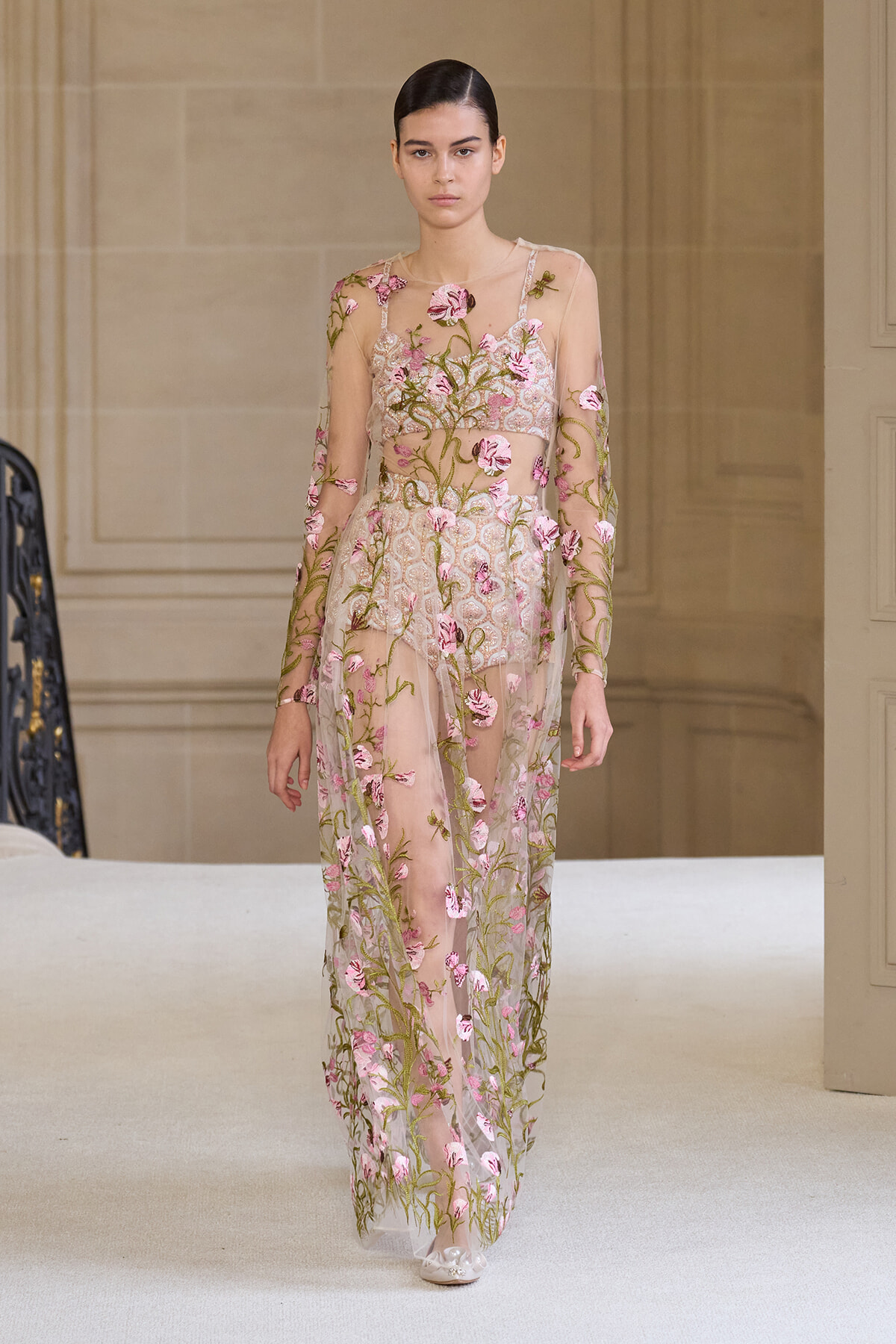 Model walking a runway in a sheer, nude-toned gown with pink floral embroidery and long sleeves.