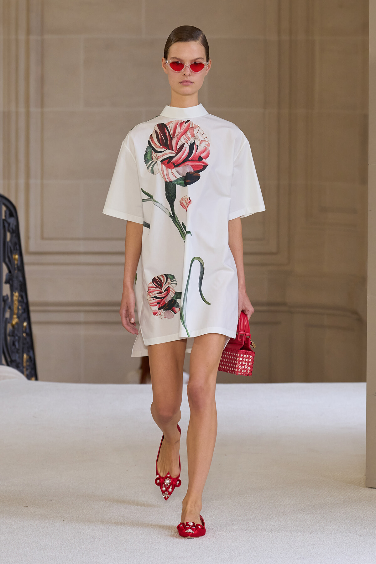Model on a runway wearing a white oversized dress with a large pink flower print, red sunglasses, red embellished flats, and a small red handbag.
