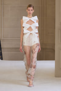Model on runway wearing a white sculptural bow-top with cutouts and sheer floral-printed pants.