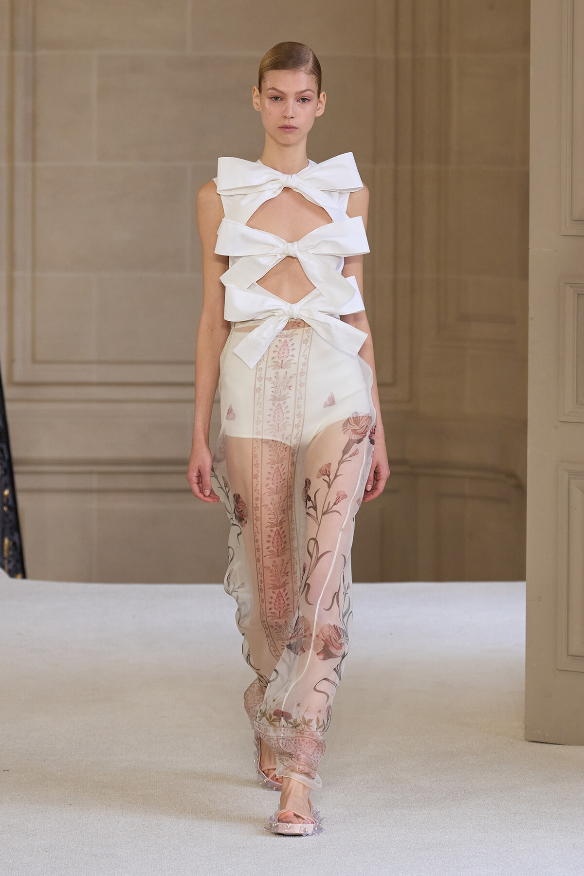 Model on runway wearing a white sculptural bow-top with cutouts and sheer floral-printed pants.