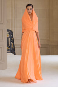 Model walking in a long peach evening gown with draped hood and twisted bodice on a runway