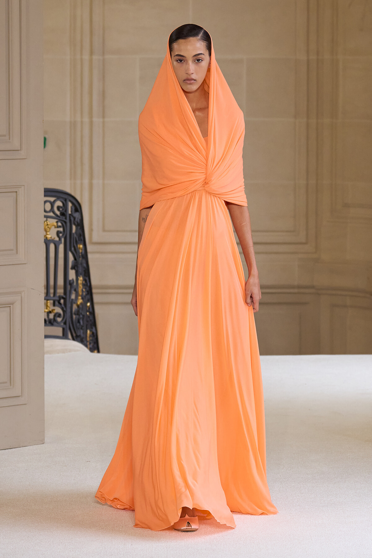 Model walking in a long peach evening gown with draped hood and twisted bodice on a runway