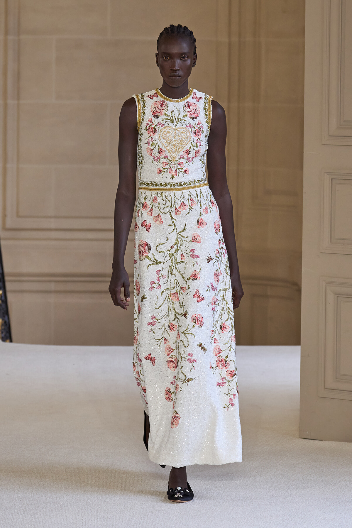 Model on a runway wearing a sleeveless white beaded maxi dress with pink floral embroidery and a heart motif at the bodice, gold trim.