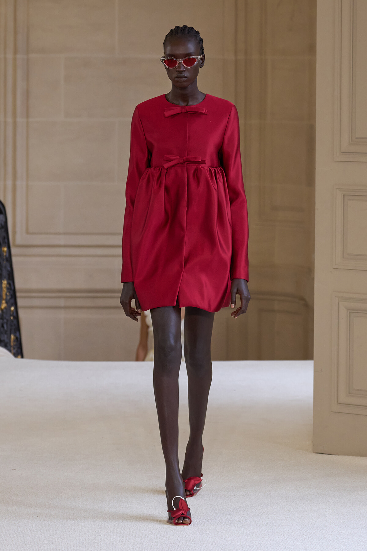 Model in a bright red, long-sleeved dress with two bow accents walking a runway, wearing red sunglasses and red heeled sandals.