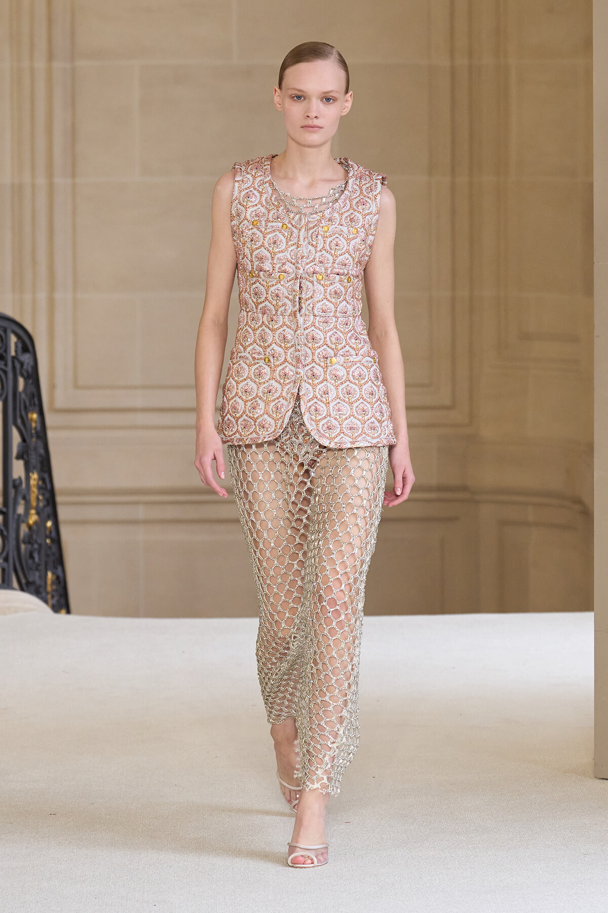 Model walking the runway in a sleeveless pink patterned vest paired with metallic mesh pants on a beige backdrop.