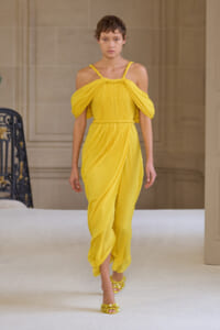 Model walking a runway in a bright yellow draped halter-style dress with off-shoulder sleeves and matching yellow sandals.
