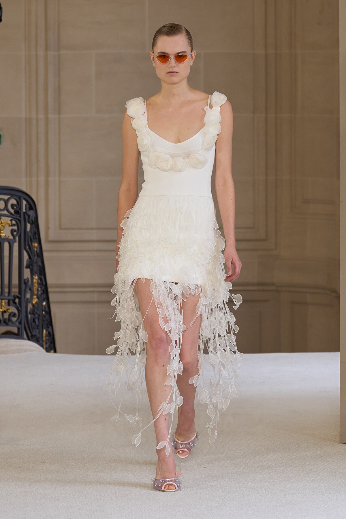 Model on a runway wearing a white sleeveless dress with flower petal appliqués and a fringed, high-low skirt, paired with orange-tinted sunglasses