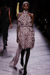 Model on a fashion show runway wearing a pink polka-dot halter dress with large front ruffles and a matching headscarf.