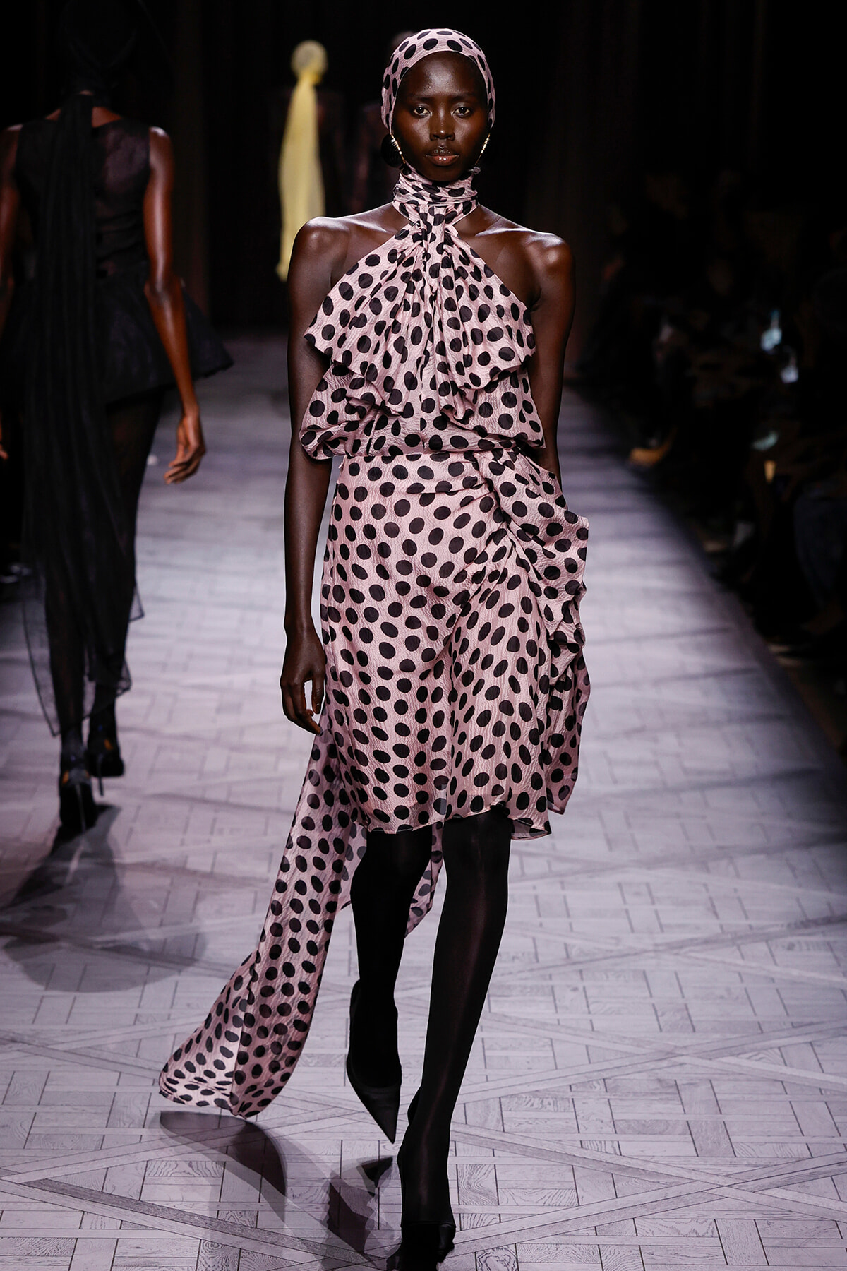 Model on a fashion show runway wearing a pink polka-dot halter dress with large front ruffles and a matching headscarf.