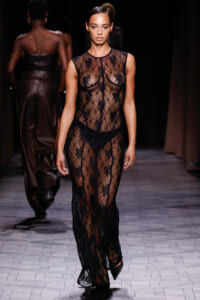 Model walking the runway in a full-length sheer black lace gown with floral pattern and black undergarments visible beneath.