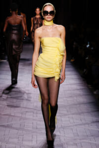 Model walks the runway in a pale yellow strapless mini dress with a cascading ruffle and sheer black tights, paired with oversized sunglasses and a headscarf.