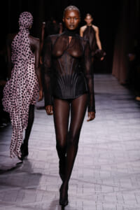 Model walks the runway in a black sheer lace bodysuit with corset panels and high neckline.