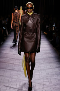 Model walks the runway in a dark brown leather double-breasted trench with a wide belt, black gloves, oversized sunglasses, and a pale yellow scarf.