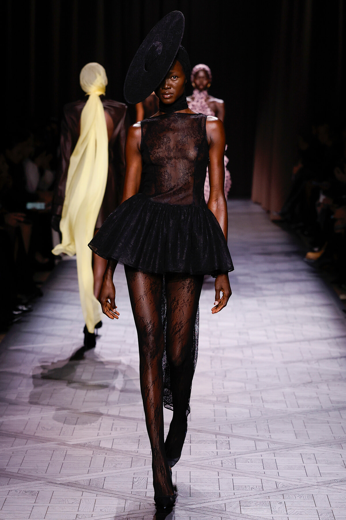 Runway model in a sheer black lace bodice with a short tutu skirt, patterned tights, and a large tilted black hat.