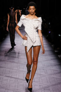Model walks a fashion show in a white textured mini dress with an oversized off-shoulder bow and black heels on a patterned runway edge sketch.