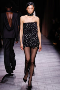 Model walks the runway in a strapless black polka-dot mini dress with cascading ruffles, paired with sheer black tights and black heels, wearing a matching polka-dot headscarf and neck scarf.
