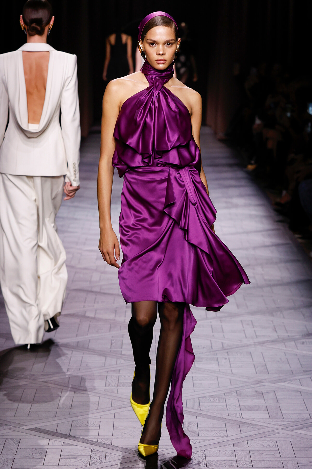 Model wearing a vivid purple satin halter dress with cascading ruffles and a wrapped sash on a runway, yellow pointed heels visible.