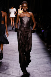 Model walks the runway in a strapless dark brown leather jumpsuit with a slim belt and oversized black bracelets, accessorized with large hoops.
