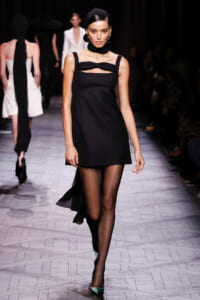 Model on a runway wearing a sleeveless black mini dress with a bow at the neckline, black tights, teal heels, and a black headband.