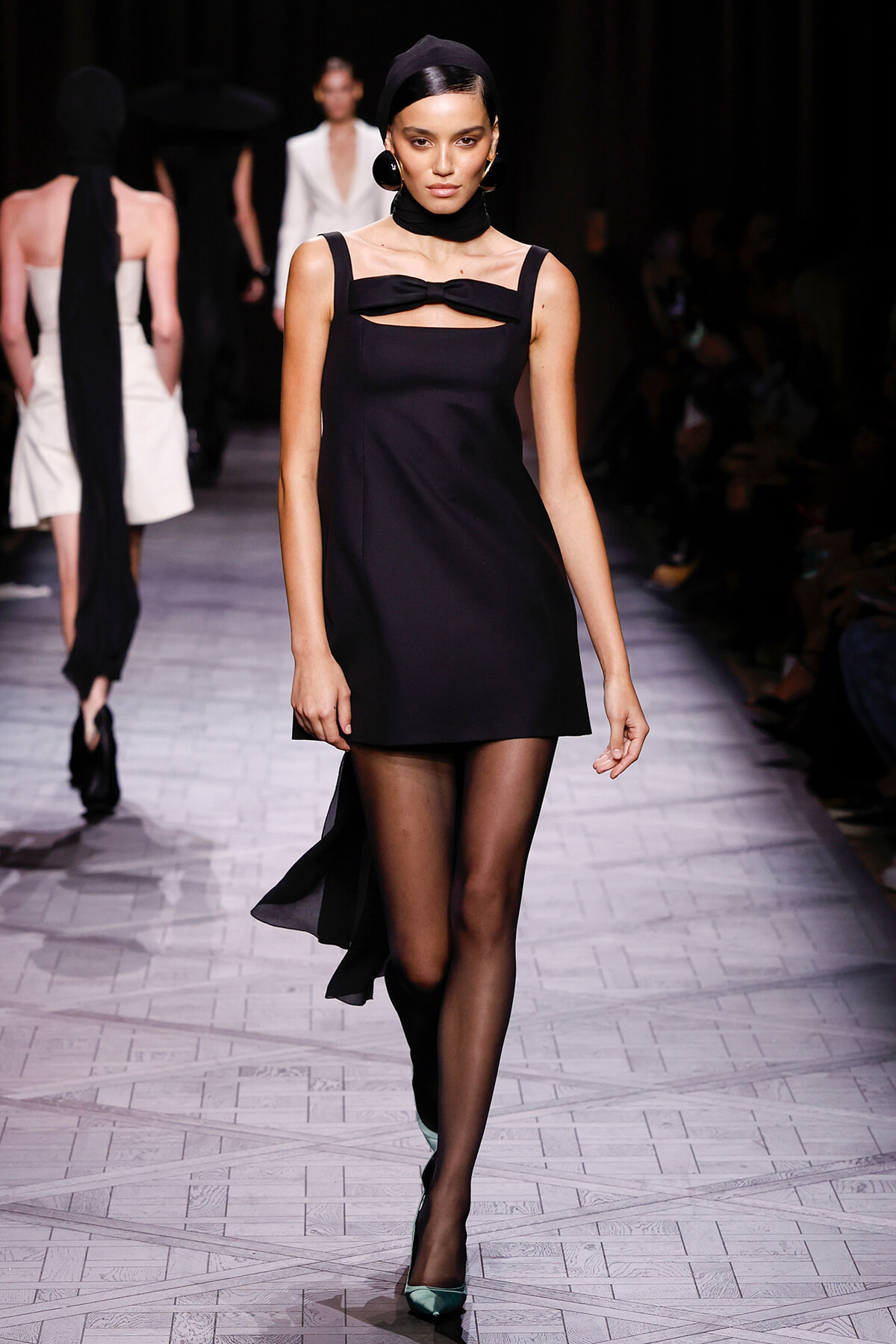 Model on a runway wearing a sleeveless black mini dress with a bow at the neckline, black tights, teal heels, and a black headband.