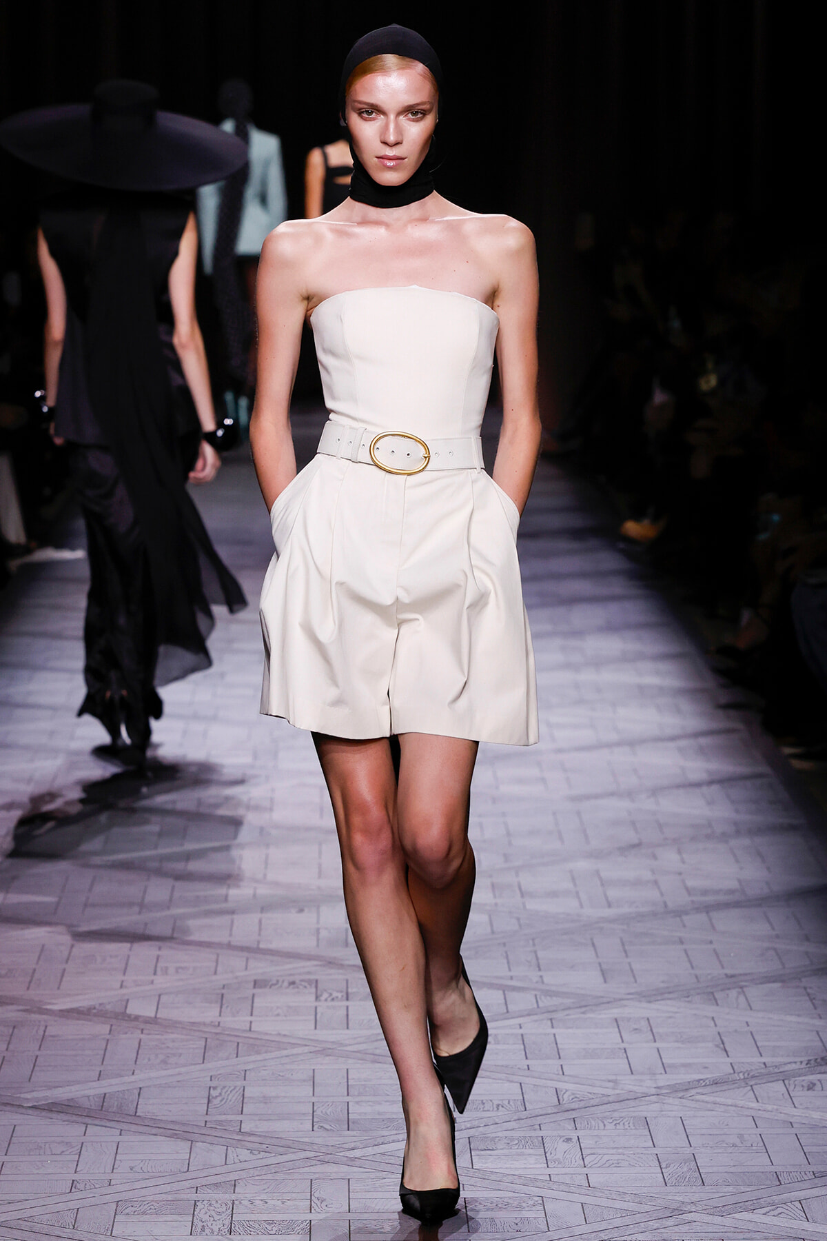 Runway model walking in a white strapless dress with a wide belt, black headscarf and heels.