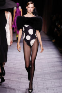 Model walking a fashion-show runway in a black fur shoulder piece over a black-and-white polka-dot bodysuit, sheer tights, and black heels.