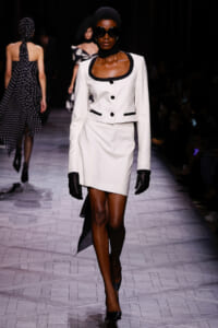 Model wearing a white tailored jacket with black trim and matching skirt, black gloves, oversized sunglasses, and a black headscarf on a runway.