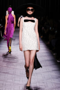 Model walks the runway in a white beaded mini dress with a black bow, oversized black hat, and dark sunglasses.
