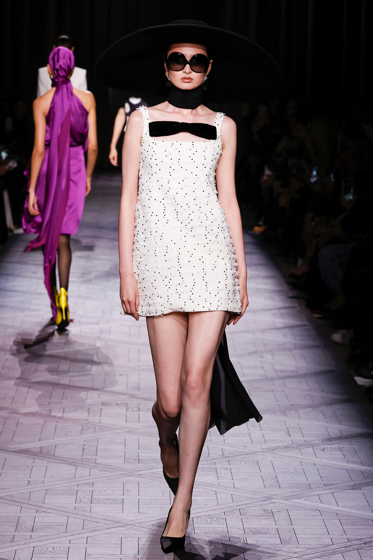 Model walks the runway in a white beaded mini dress with a black bow, oversized black hat, and dark sunglasses.