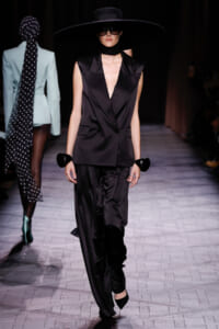 Model on a runway wearing a black satin sleeveless blazer with a deep V, matching wide-leg pants, oversized brim hat and sunglasses.