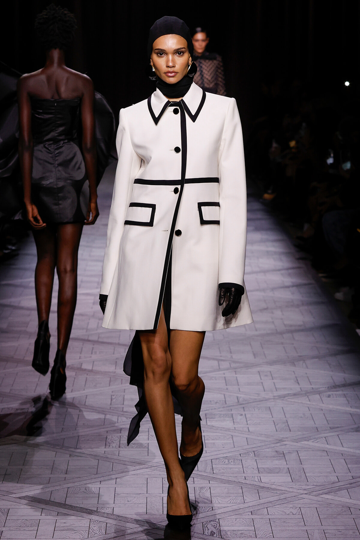 Model walking a fashion runway in a white tailored coat with black piping and pockets, paired with a black headscarf and gloves.