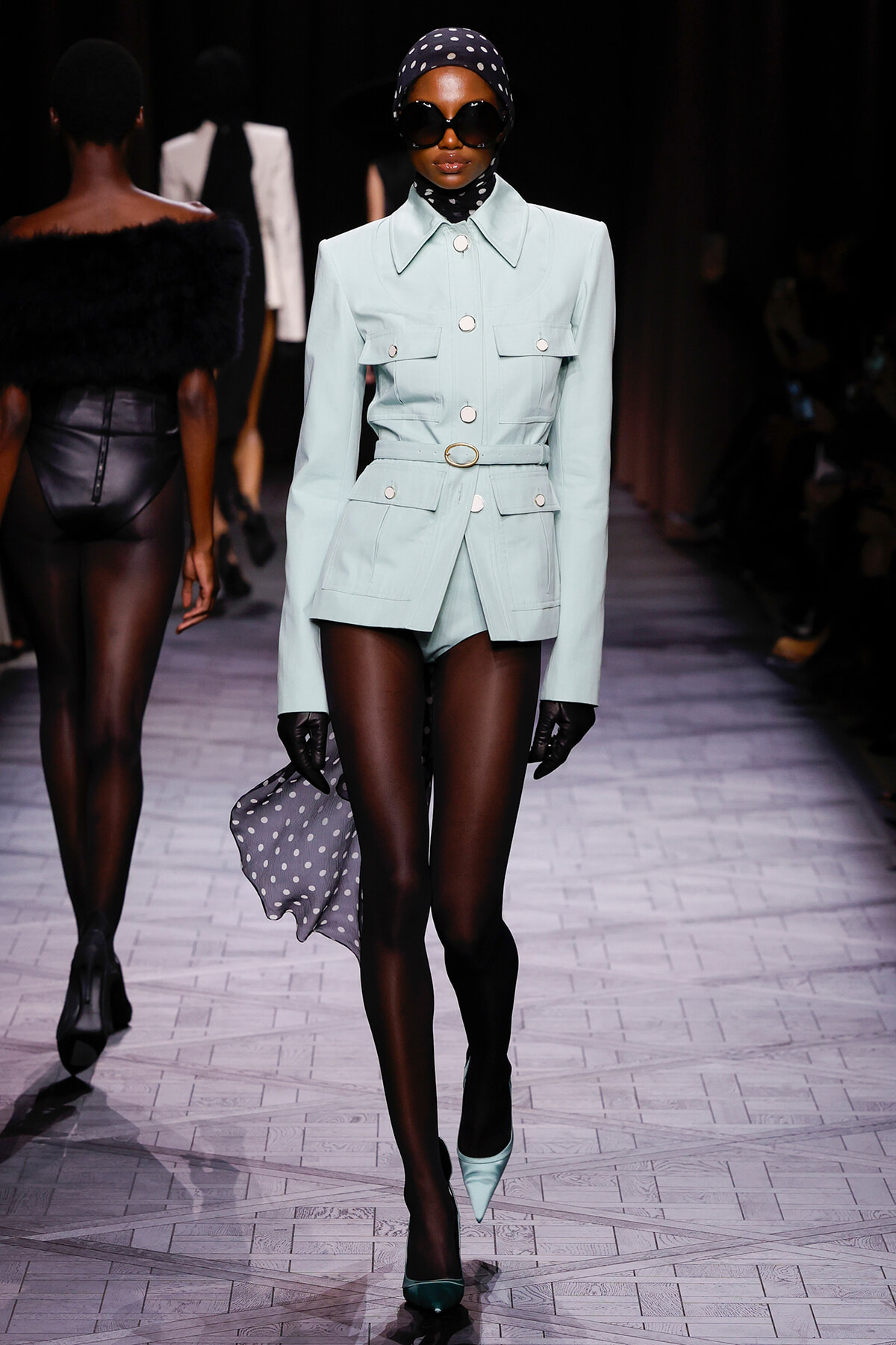 Model on a runway in a pale aqua tailored blazer with matching shorts, belt, black tights, mint heels, oversized sunglasses, and a navy polka-dot headscarf holding a matching scarf.