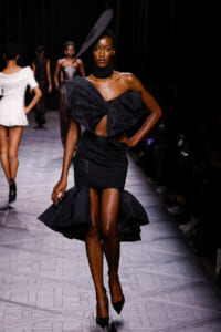 Model walks the runway in a black sculptural dress with a large bow and oversized hat, posing confidently.