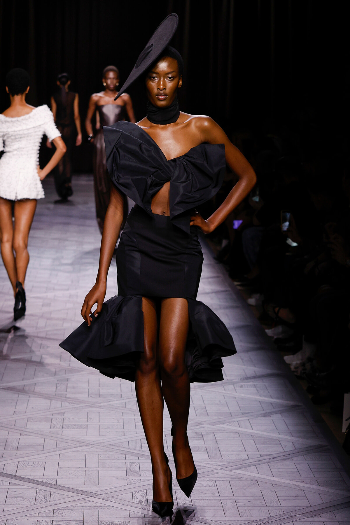 Model walks the runway in a black sculptural dress with a large bow and oversized hat, posing confidently.