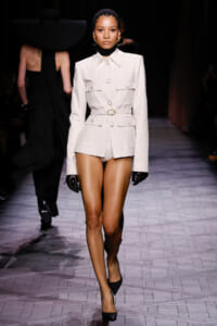 Model walks runway wearing an ivory tailored belted blazer with matching shorts, black gloves, and black heels; headband and gold earrings complete look.