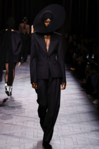Model on a runway wearing a black tailored blazer with a plunging neckline and oversized circular black hat obscuring the face.