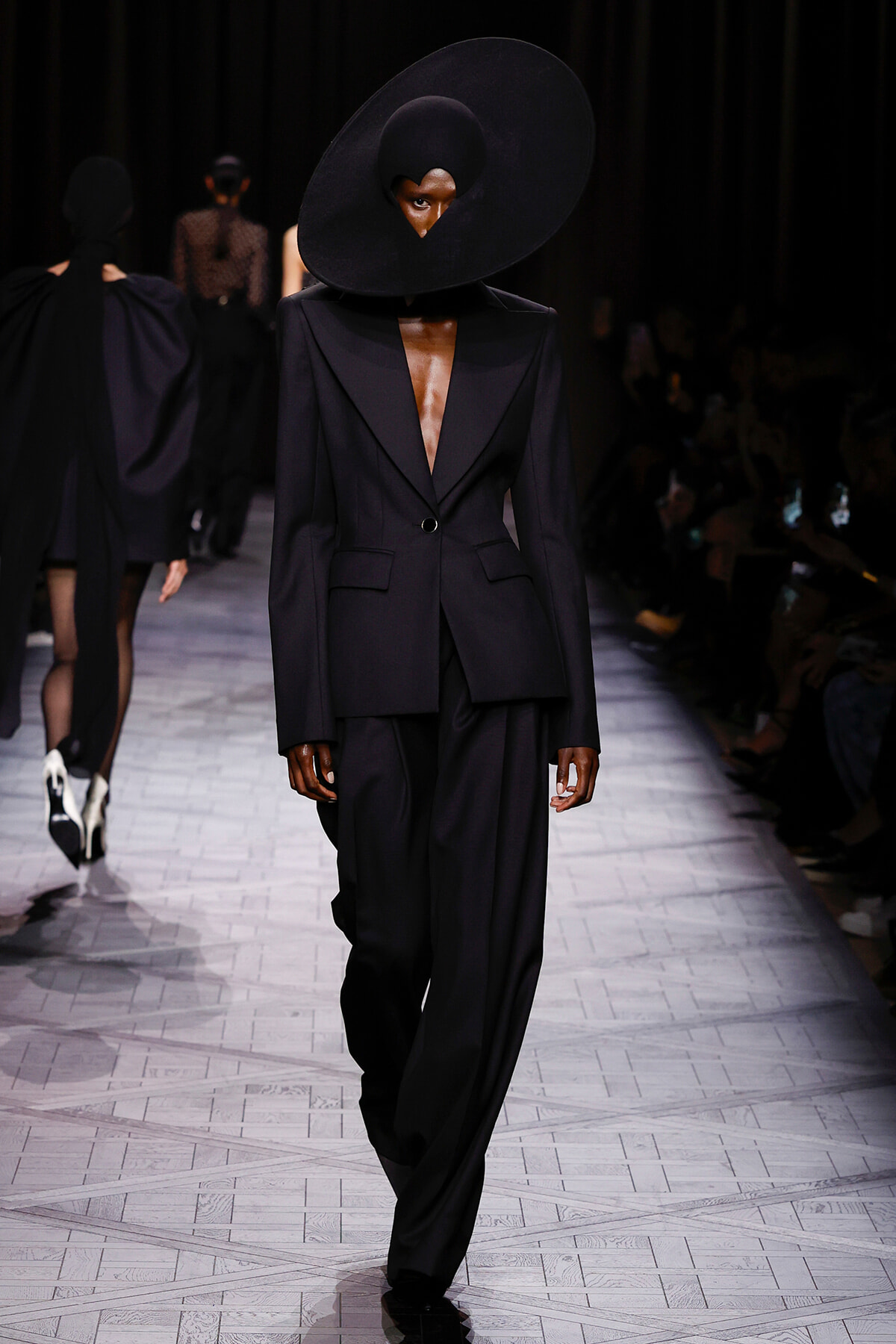 Model on a runway wearing a black tailored blazer with a plunging neckline and oversized circular black hat obscuring the face.