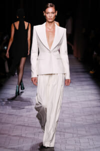 Model walks a fashion runway wearing a white tailored blazer with a deep V-neck and matching wide-leg trousers, accessorized with statement earrings.