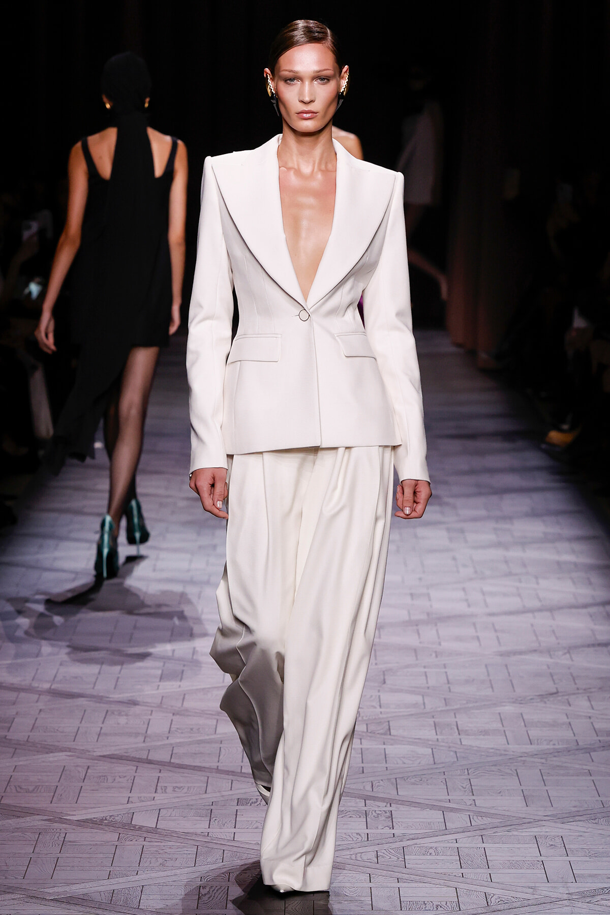 Model walks a fashion runway wearing a white tailored blazer with a deep V-neck and matching wide-leg trousers, accessorized with statement earrings.