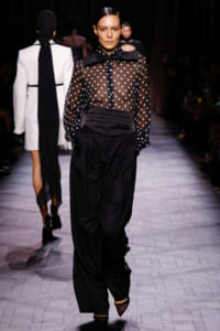 Model walks a fashion runway in a sheer black polka-dot blouse with a large bow at the neck and high-waisted, draped black trousers.