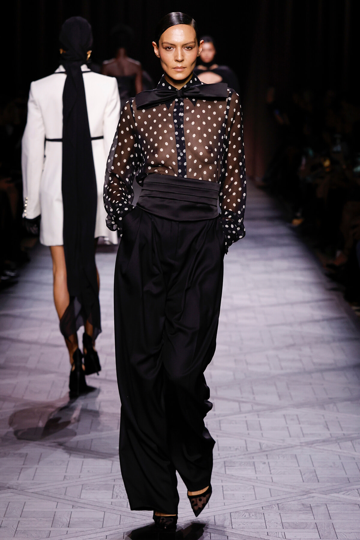 Model walks a fashion runway in a sheer black polka-dot blouse with a large bow at the neck and high-waisted, draped black trousers.