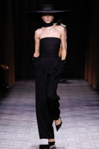 Model walking a runway in a strapless black jumpsuit with wide-leg pants, oversized black hat, and black gloves.