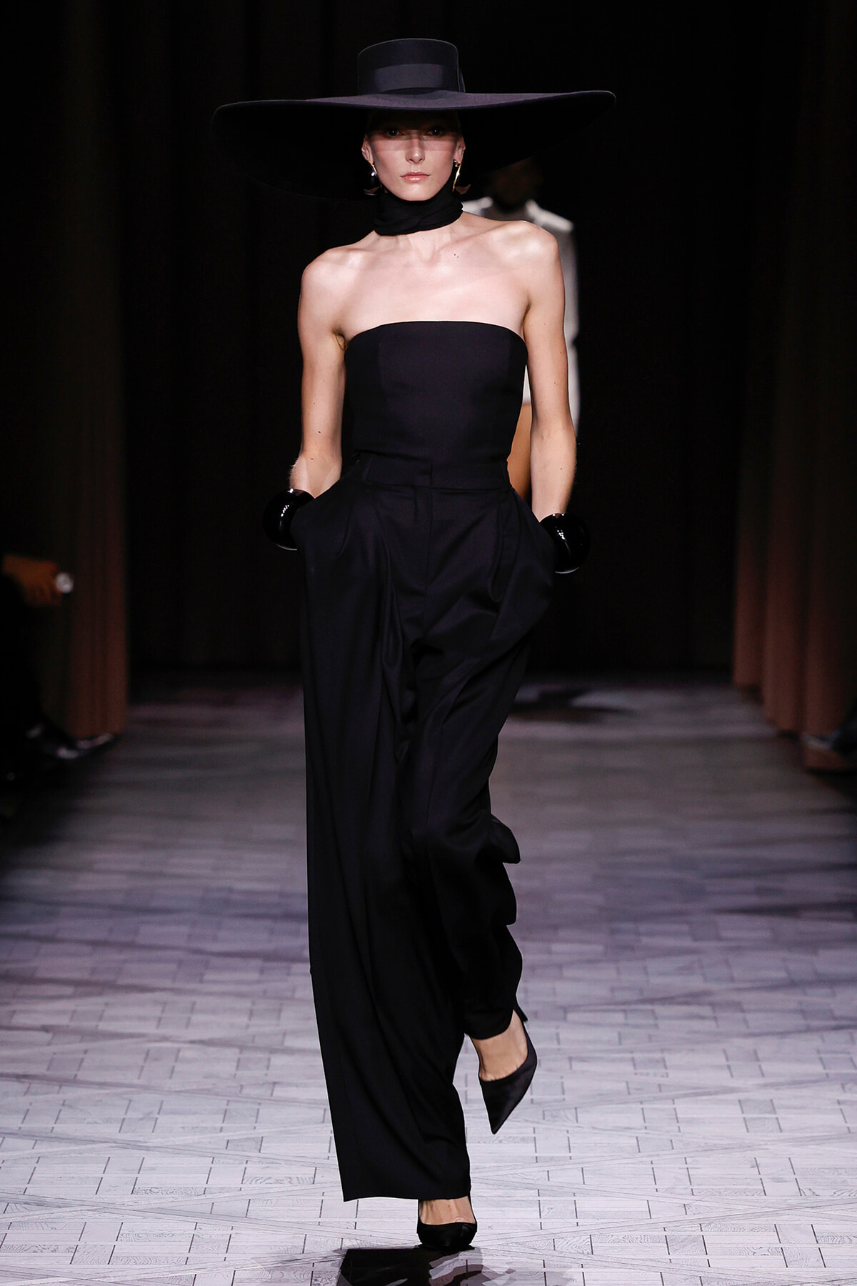 Model walking a runway in a strapless black jumpsuit with wide-leg pants, oversized black hat, and black gloves.