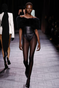 Model walks the runway in an off-the-shoulder black fur stole over a sleek black leather bodysuit and sheer tights with pointed heels.