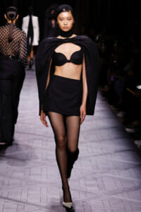 Model walks the runway in an all-black ensemble: cape draped over shoulders, black bra, high-waisted mini skirt, sheer tights, and white heels during a fashion show.