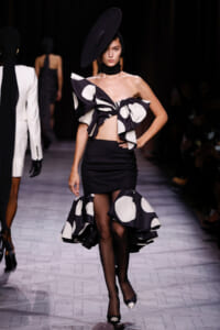 Model on a runway wearing a black and white polka-dot off-shoulder bow top with a black mini skirt and large ruffled hem; wide black hat and tights.