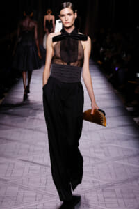 Runway model in a sheer black top with a large bow t-strap, high-waisted belt, and wide black pants, carrying a curved metallic clutch.