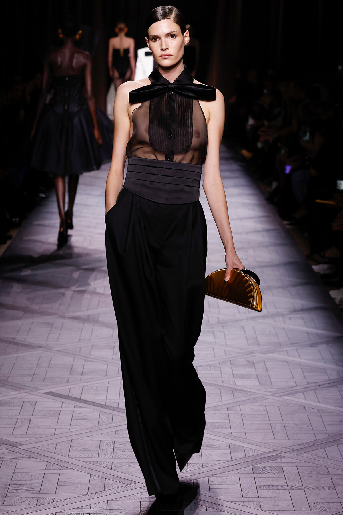 Runway model in a sheer black top with a large bow t-strap, high-waisted belt, and wide black pants, carrying a curved metallic clutch.