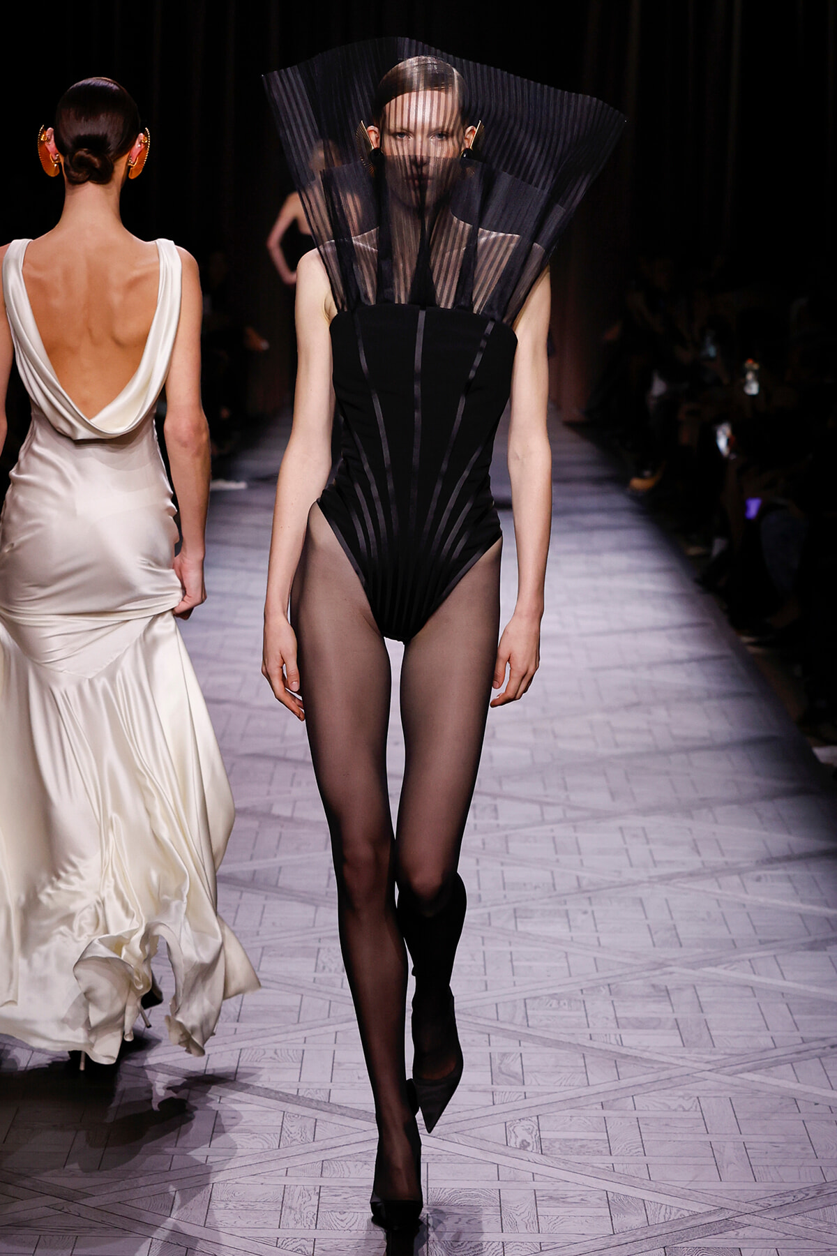 Model walks the runway in a black sculptural bodysuit with a tall pleated sheer collar and sheer tights.