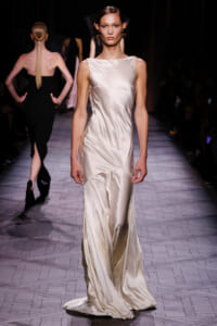Model walks the runway in a sleeveless ivory satin gown with a fitted bodice and flowing, draped skirt.
